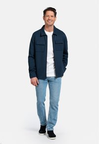Marine lichtgewicht jack met twee borstzakken, gedragen over een wit T-shirt en lichtblauwe jeans. Model staat voor een neutrale achtergrond.