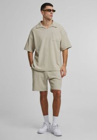 Beige oversized polo shirt met een borstzak, gecombineerd met bijpassende shorts. De outfit heeft een gestructureerde stof en witte sneakers.