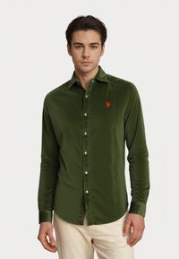 Giovane uomo che indossa una camicia verde oliva con bottoni e un logo rosso sul petto, e pantaloni beige chiaro, in piedi contro uno sfondo bianco.