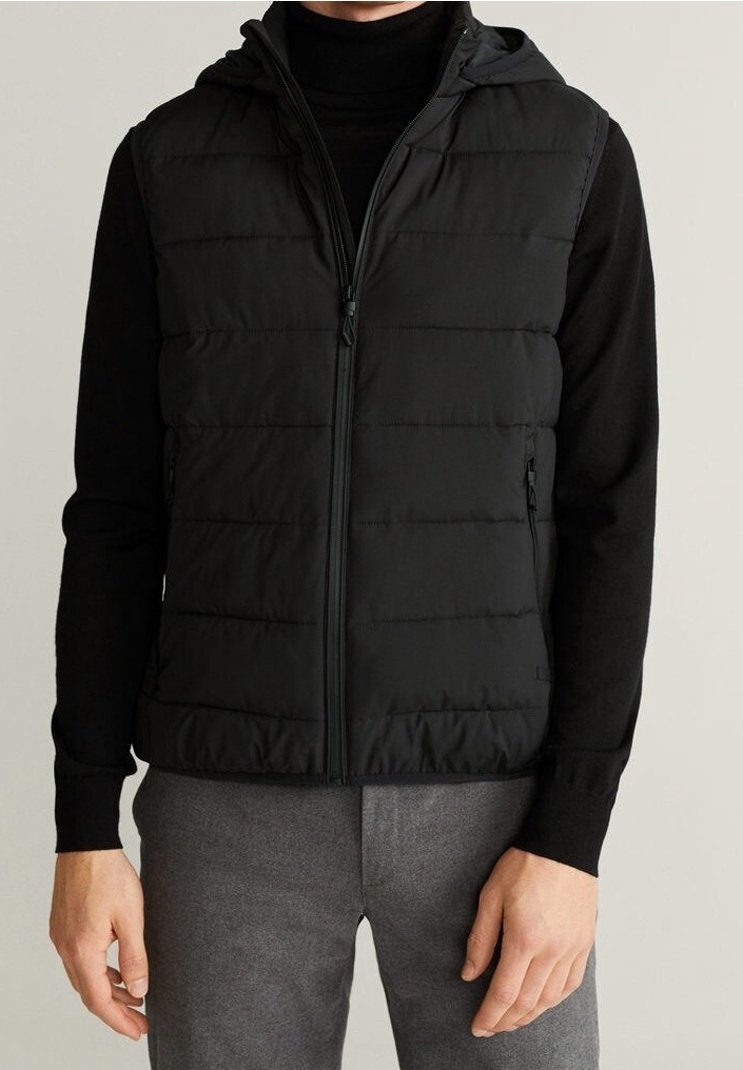 Homme portant une veste noire matelassée sans manches avec fermeture éclair, par-dessus un haut noir à manches longues et un pantalon gris, debout, les bras détendus le long du corps.