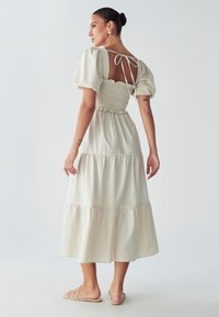 Grietos spalvos midi suknelė su pūkuotomis rankovėmis, sukurtu liemeniu ir sluoksniuotu sijonu. Yra raištelių uždarymas nugaroje ir minkšta, tekstūruota medžiaga.