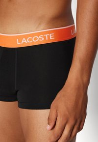 Black cotton boxer shorts with an orange waistband featuring white "LACOSTE" text. Close-up of waistband and fabric texture.