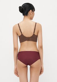 Reggiseno marrone con spalline regolabili e chiusura a gancio; abbinato a un bikini color bordeaux con una texture liscia e un design a vita media.