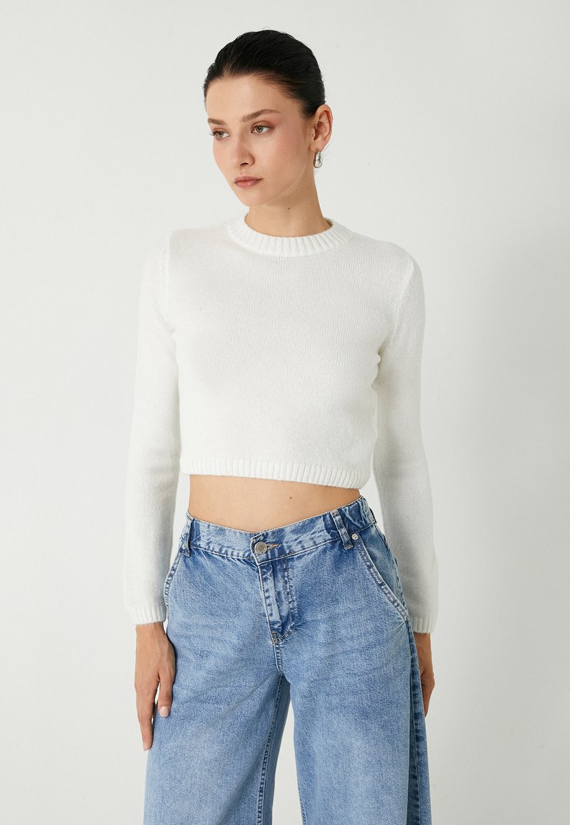 Witte cropped trui met lange mouwen, geribbelde halslijn en zoom, gecombineerd met lichtblauwe high-waisted wijde jeans met knoopsluiting.