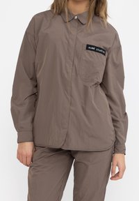 Žena v volném hnědém tričku s dlouhými rukávy a odpovídajících kalhotách, na kapse trička je štítek "esmé studios," stojí na bílém pozadí.