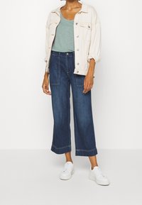 Ljus crème denimjacka över en grön topp, parat med vida mörkblå jeans och vita sneakers. Slät tyg med enkel design.