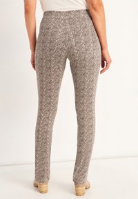 Pantalons beiges ajustés et texturés avec un motif subtil, dotés d'une taille mi-haute et de détails de couture au dos. Portés avec des talons de couleur claire.