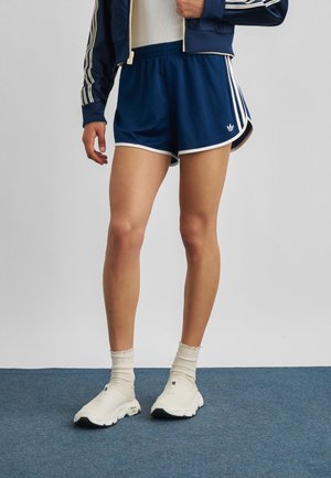 Person iført navyblå atletikshorts og matchende jakke med hvide sokker og lyse slip-on sneakers, der står på blå tæppe.