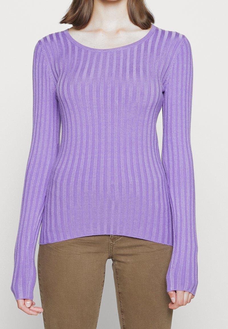 Femme portant un pull ajusté à manches longues en maille côtelée lavande, associé à un pantalon marron, debout devant un fond uni.