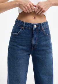 Jean en denim bleu foncé taille haute avec fermeture à boutons et coutures visibles ; comprend des poches avant et une coupe légèrement décontractée.