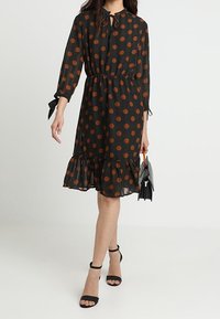 Černé šaty s puntíky a volánkovým lemem, dlouhé rukávy s detailem na zavazování a staženým pasem. V kombinaci s černými sandály na podpatku a kabelkou.