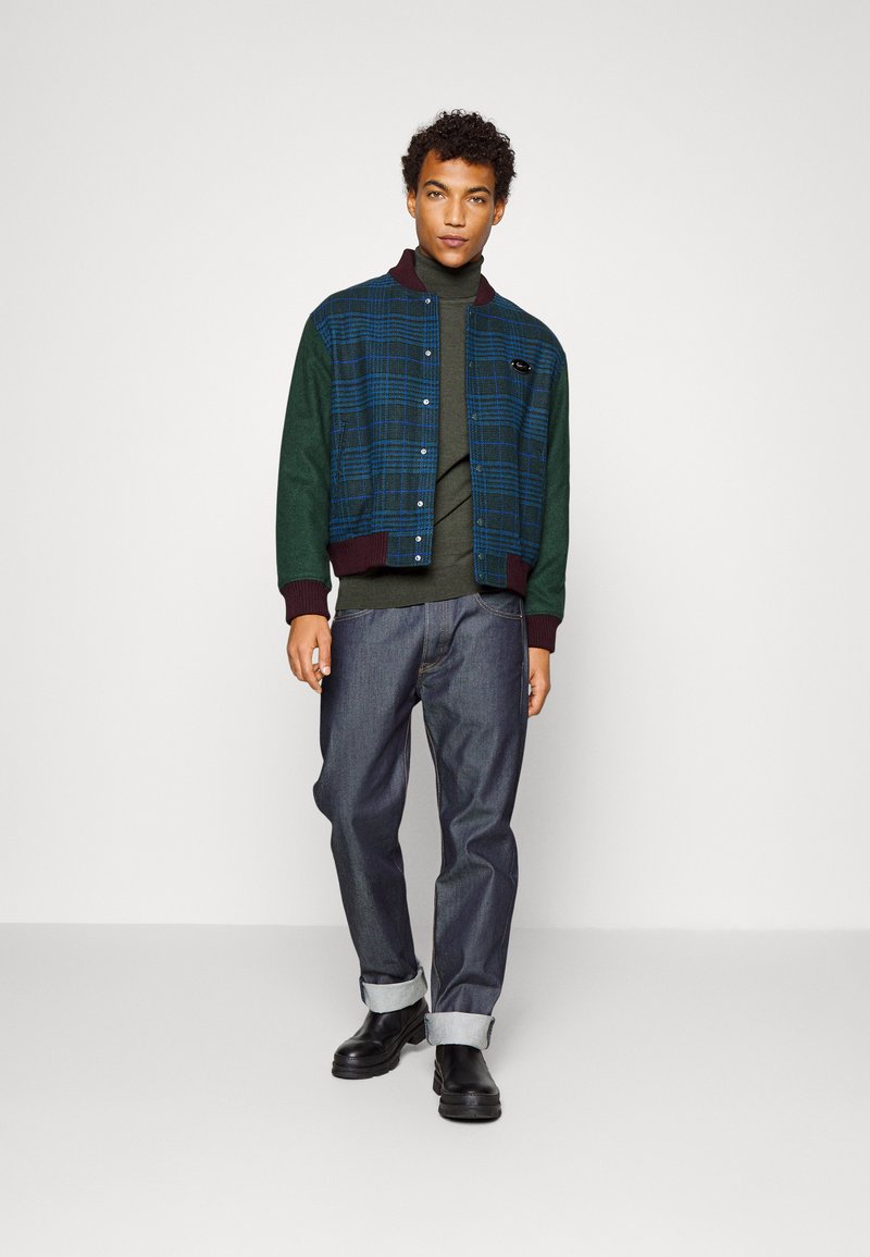 Veste bomber à carreaux en vert et bleu avec des poignets côtelés, portée sur un turtleneck sombre et associée à un jean en denim foncé retroussé et des bottes noires.