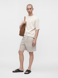 Man wearing light beige shorts, cream short-sleeve sweater, black sandals, and carrying a brown leather shoulder bag against a plain background.