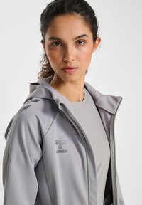 Ljusgrå zip-up jacka med huva, med en mjuk textur, diskret logo på bröstet och kontrasterande detaljer på axlarna.