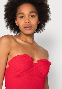 Top sem alças vermelho com decote em camadas, design justo, tecido texturizado e detalhes de costura. A modelo tem cabelo encaracolado natural e maquilhagem neutra.