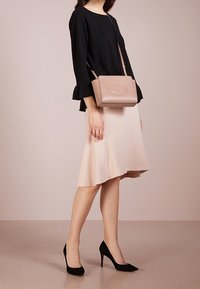 Světle růžová kožená crossbody taška s embosovaným logem, obdélníkový tvar, nastavitelné rameno, v kombinaci s černým topem, béžovou sukní a černými podpatky.