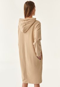 Vestido largo beige con capucha y un corte relajado, hecho de material suave. Presenta bolsillos y una costura en la parte posterior. Diseño simple, sin patrones.
