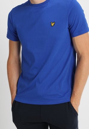 Homme portant un t-shirt bleu uni à manches courtes avec un petit logo d'oiseau jaune sur le côté gauche de la poitrine et un pantalon noir, les mains dans les poches.