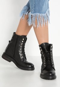 Bottes courtes en cuir noir avec devant à lacets, œillets argentés et fermeture éclair. Surface texturée et détail de talon en bois.