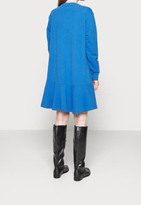 Sininen hupparimekko pitkillä hihoilla ja levenevällä helmalla. Yhdistetty kiiltäväpintaisten mustien polvipitkien saappaidensa kanssa, joissa on krokokoski.