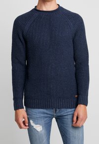 Granatowy sweter dziany z teksturowanym wzorem, ściągaczami na mankietach i u dołu. Posiada rękawy raglanowe i okrągły dekolt. Noszony z postrzępionymi dżinsami.