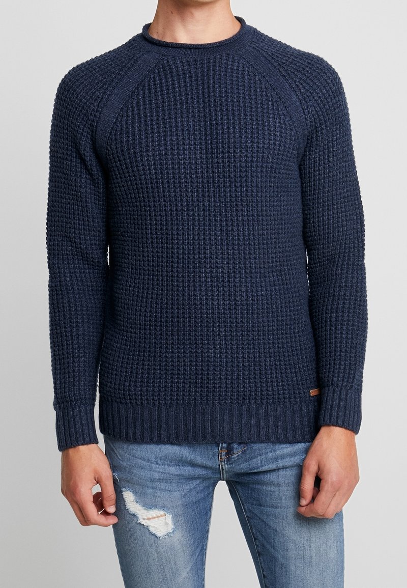 Granatowy sweter dziany z teksturowanym wzorem, ściągaczami na mankietach i u dołu. Posiada rękawy raglanowe i okrągły dekolt. Noszony z postrzępionymi dżinsami.