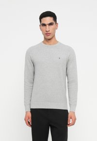 Νεαρός άνδρας φορώντας ένα ανοιχτό γκρι ριγωτό πουλόβερ και μαύρο παντελόνι, κοιτάζοντας μπροστά σε έναν απλό λευ�κό φόντο.