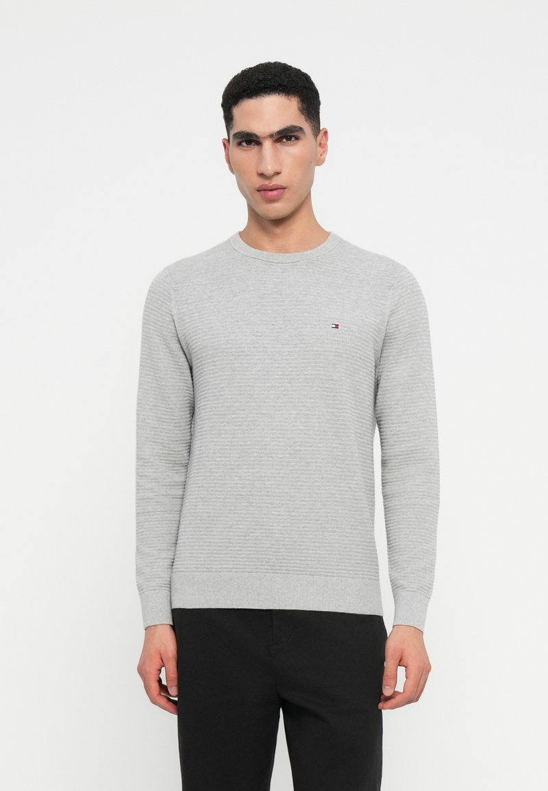 Νεαρός άνδρας φορώντας ένα ανοιχτό γκρι ριγωτό πουλόβερ και μαύρο παντελόνι, κοιτάζοντας μπροστά σε έναν απλό λευκό φόντο.