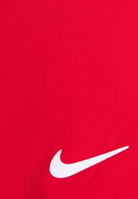 Logo Nike swoosh blanc sur fond de tissu rouge vif, visible dans le coin inférieur droit.