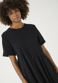 Juoda suknelė su apvaliu iškirpimu ir trumpomis sukniotomis rankovėmis. Yra priglausdanti liemenė ir plačiai krentanti, surinkta sijoninė dalis. Minkšta medžiagos tekstūra.