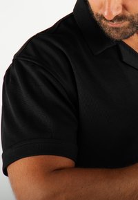 Polo noir à manches courtes, en tissu texturé, avec un col classique, présentant des coutures discrètes le long des coutures.