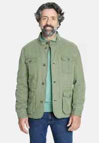 Hombre con cabello rizado y barba, vistiendo una chaqueta verde claro sobre un suéter teal y jeans azules, sonriendo a la cámara.