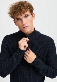 Dunkelblaues, zip-up Sweatshirt mit gerippter Textur, hohem Kragen und langen Ärmeln. Das Modell greift mit beiden Händen den Reißverschluss.