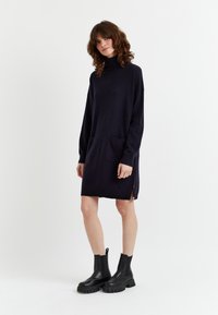 Abito a maglia blu navy con colletto alto, maniche lunghe, due tasche frontali, spacchi laterali e un piccolo dettaglio rosso. Indossato con stivali neri chunky.