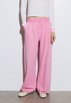 Personne portant un pantalon rose taille haute à jambes larges avec une ceinture élastique et des chaussures blanches, debout devant un fond blanc uni.