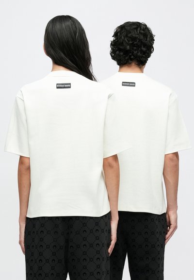 Short-sleeve, cream cotton t-shirts with a relaxed fit, featuring a black label at the back. Worn with patterned dark pants.