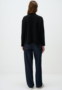 Zwarte sweatshirt in een ontspannen pasvorm met een ronde hals, voorzien van een blauw accentpatroon op de linkermouw. Gecombineerd met losse blauwe jeans.