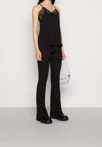 Camisole noire avec bordure en dentelle, associée à un pantalon noir évasé. Un sac à main violet clair complète la tenue. Matériaux texturés visibles.