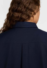 Camisa azul marinho com colarinho estruturado, pregas nas costas e tecido texturizado. O design é simples e discreto, focando na funcionalidade.