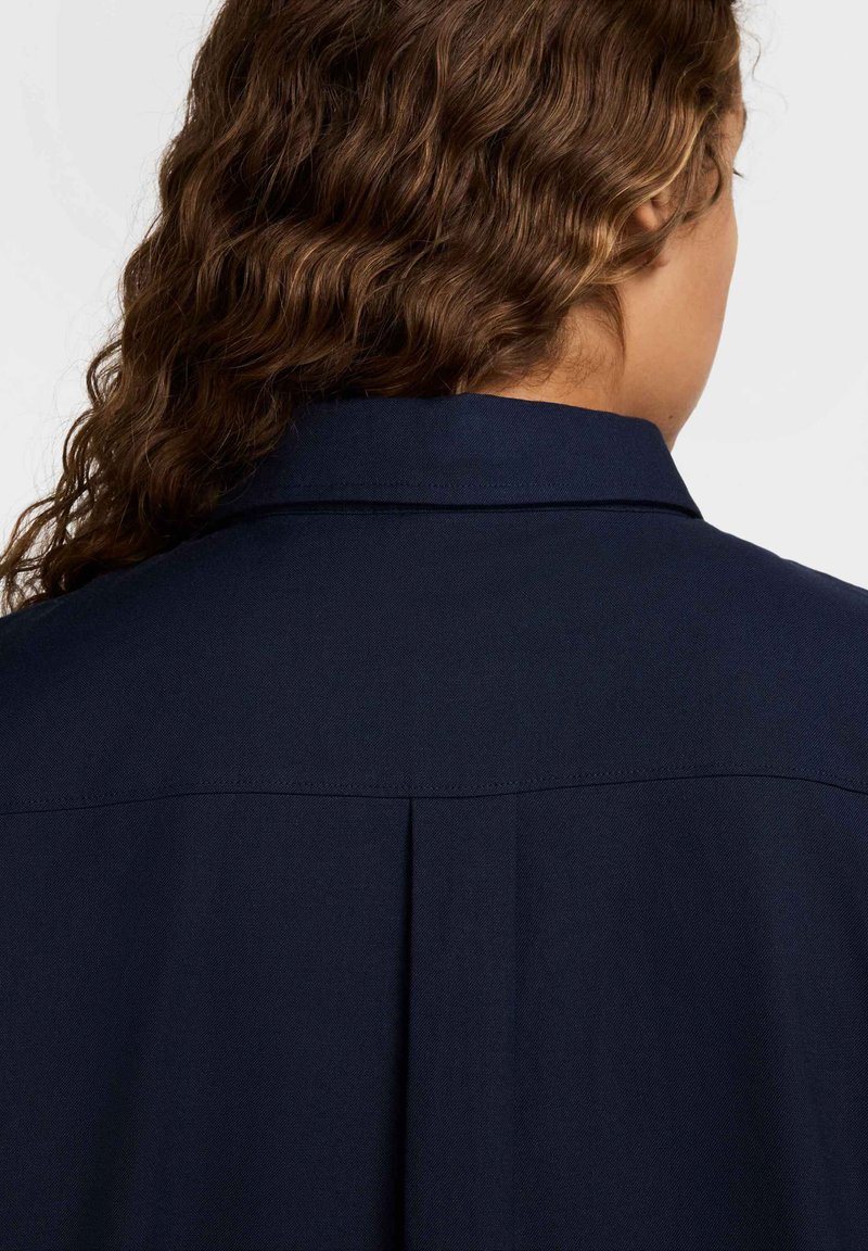 Chemise bleu marine avec un col structuré, une pli au dos et un tissu texturé. Le design est simple et discret, mettant l'accent sur la fonctionnalité.