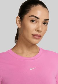 Donna con i capelli scuri raccolti, che indossa una t-shirt Nike rosa con un piccolo logo swoosh bianco su uno sfondo grigio uniforme.