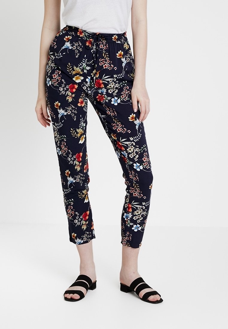Femme portant un pantalon bleu marine évasé avec un imprimé floral multicolore, un haut blanc et des sandales noires à bout ouvert, debout devant un fond uni.