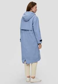 Lichtblauwe waterdichte regenjas met een capuchon, voorzien van een cropped bovenlaag, zwarte accenten en een ventilerende achterkant. Draagt over crèmekleurige broek en witte schoenen.