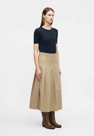 Γυναίκα όρθια με μπλε μπλούζα κοντομάνικη, μπεζ πλισέ μεσάτο φούστα και καφέ μπότες μέχρι  τη μέση της γάμπας, σε απλό φωτεινό φόντο.