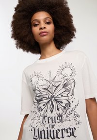 Biała, oversize'owa koszulka z krótkim rękawem, z grafiką czarnej motyli, motywami słońca i kwiatowymi akcentami. Okrągły dekolt.