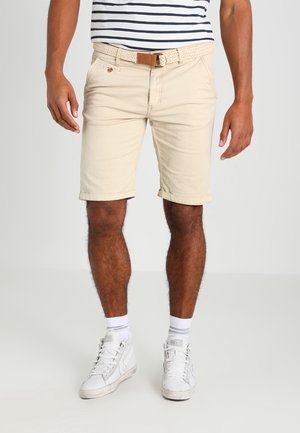 Homme portant un short beige avec une ceinture tressée, des baskets montantes blanches, des chaussettes blanches et une chemise rayée raccourcie au-dessus de la taille.