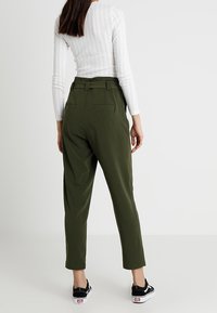 Pantalones verde oliva, ajustados, de cintura alta, con bolsillos laterales y un diseño de pierna estrecha; combinados con zapatos negros y una blusa de rayas blancas.