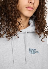 Sudadera con capucha gris que presenta el texto "ELEMENT SKATEBOARDS & MORE" en bordado azul oscuro, cordones y un tejido suave y texturizado.