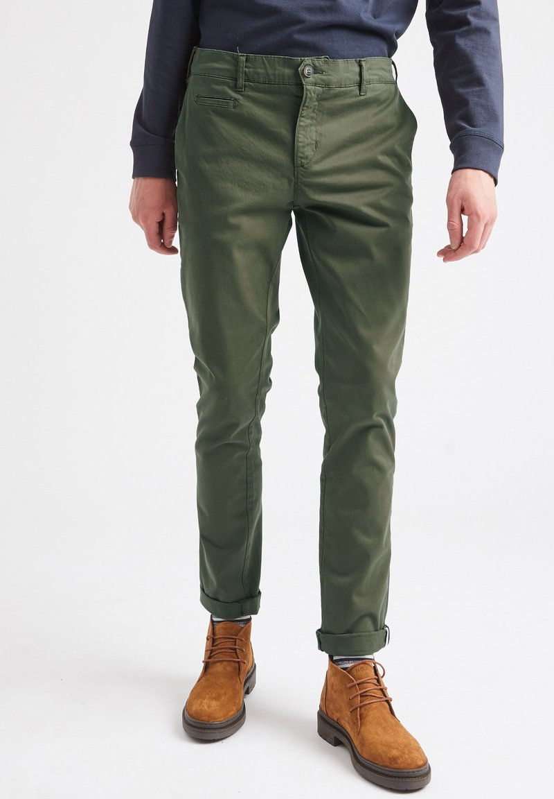 Chinos verdes de corte slim, con una textura suave, que cuentan con una cremallera, bolsillos frontales y dobladillos enrollados, combinados con botas de ante marrón hasta el tobillo.