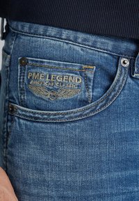 Jeans in denim blu con una texture sbiadita, dotati di un'etichetta con il logo "PME LEGEND AMERICAN CLASSIC" e cuciture gialle a contrasto.